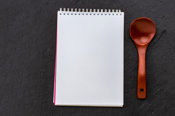 Notepad for your recipe with wooden spoon over black stone background. Top view with copy space