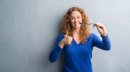 Young redhead woman over grey grunge wall holding optical glasses happy with big smile doing ok sign, thumb up with fingers, excellent sign