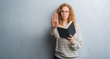 Young redhead woman over grey grunge wall reading a book with open hand doing stop sign with...