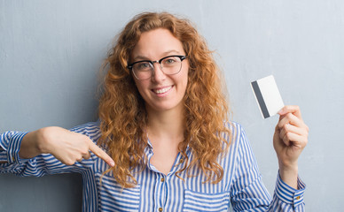 Young redhead woman over grey grunge wall holding credit card with surprise face pointing finger to...