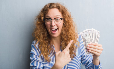 Young redhead woman over grey grunge wall holding dollars very happy pointing with hand and finger