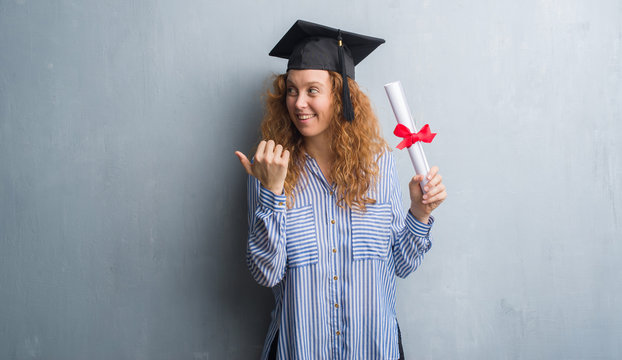 Young redhead graduated woman over grey grunge wall holding diploma pointing and showing with thumb up to the side with happy face smiling