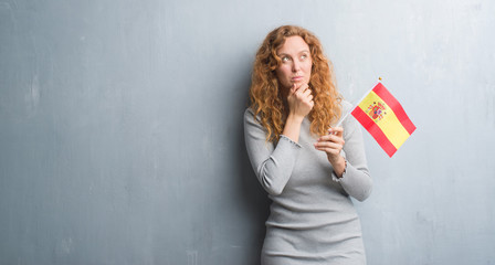 Young redhead woman over grey grunge wall holding flag of Spain serious face thinking about question, very confused idea