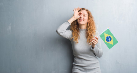 Young redhead woman over grey grunge wall holding flag of Brazil with happy face smiling doing ok sign with hand on eye looking through fingers