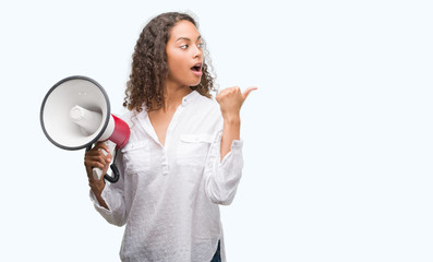 Young hispanic woman holding megaphone pointing and showing with thumb up to the side with happy...