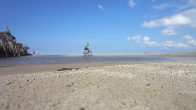 A man rides a bike through a sandy beach next to an old wooden pier