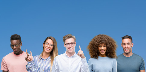 Composition of group of friends over blue blackground showing and pointing up with finger number one while smiling confident and happy.