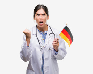 Young hispanic doctor woman holding flag of germany annoyed and frustrated shouting with anger, crazy and yelling with raised hand, anger concept