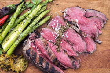 Traditionell gegrilltes dry aged Roastbeef Rindersteak aufgeschnitten mit grünen Spargel und Tomaten als closeup auf einem Holzbrett 
