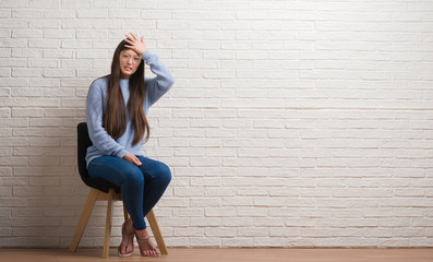 Young Chinese woman sitting on chair over brick wall stressed with hand on head, shocked with shame and surprise face, angry and frustrated. Fear and upset for mistake.