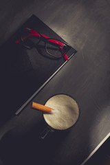 Cup of cappuccino garnished with cinnamon sticks on a dark office desk with calendar, pencil, glasses and cellphone