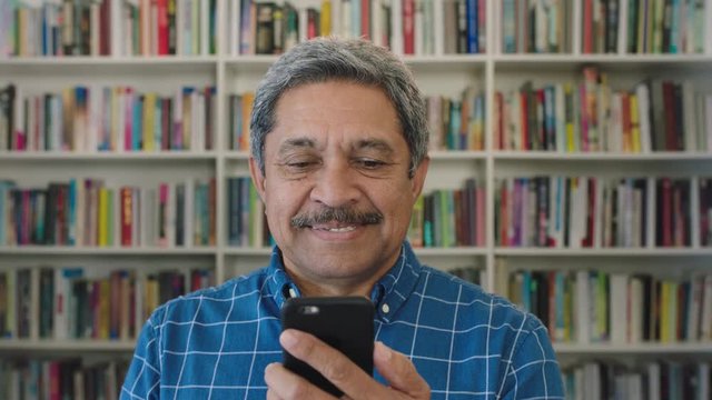 close up portrait of happy mature hispanic man texting browsing using smartphone mobile technology enjoying digital communication in library bookshelf background