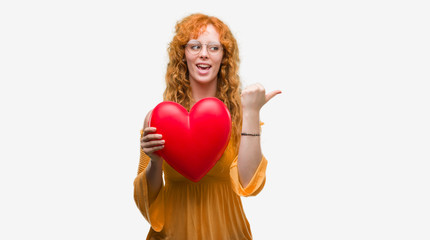 Young redhead woman in love holding red heart pointing and showing with thumb up to the side with happy face smiling