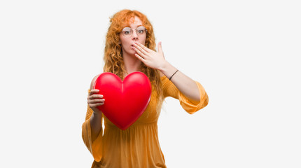 Young redhead woman in love holding red heart cover mouth with hand shocked with shame for mistake, expression of fear, scared in silence, secret concept