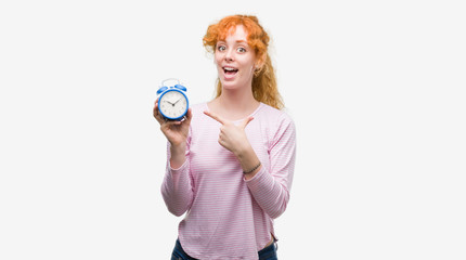 Young redhead woman holding alarm clock very happy pointing with hand and finger