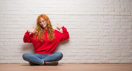Young redhead woman sitting over brick wall looking confident with smile on face, pointing oneself with fingers proud and happy.