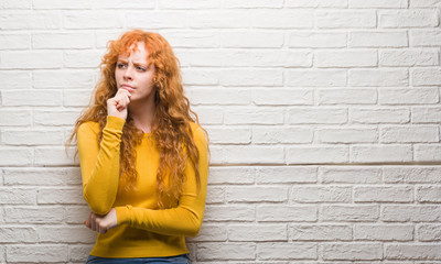 Young redhead woman standing over brick wall serious face thinking about question, very confused...