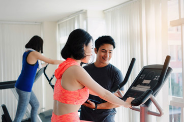 Fototapeta na wymiar asian young woman slim body exercising on bicycle machine with young man personal trainer in fitness gym, bodybuilder, healthy lifestyle, fitness, workout and sport training concept