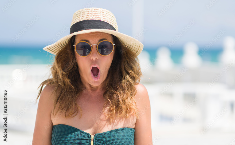 Wall mural middle age brunette woman wearing sunglasses by the pool scared in shock with a surprise face, afrai