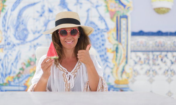 Middle age brunette woman sitting at the table eating watermelon happy with big smile doing ok sign, thumb up with fingers, excellent sign
