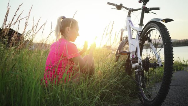 Happy woman after riding bicycle looking into smartphone, road, river at sunset outdoors background