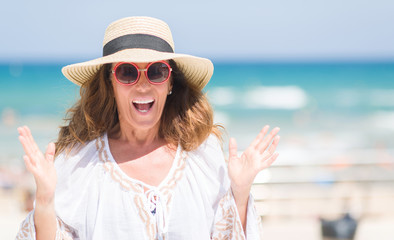Middle age brunette woman wearing summer hat on vacations by the beach very happy and excited,...