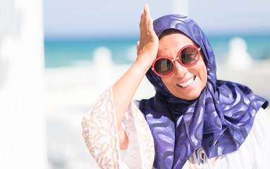 Middle age brunette arabian woman by the pool wearing sunglasses stressed with hand on head, shocked with shame and surprise face, angry and frustrated. Fear and upset for mistake.