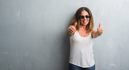 Middle age hispanic woman over grey wall wearing sunglasses approving doing positive gesture with hand, thumbs up smiling and happy for success. Looking at the camera, winner gesture.