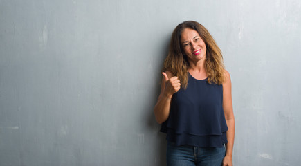 Fototapeta premium Middle age hispanic woman standing over grey grunge wall doing happy thumbs up gesture with hand. Approving expression looking at the camera with showing success.
