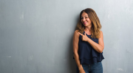 Fototapeta na wymiar Middle age hispanic woman standing over grey grunge wall cheerful with a smile of face pointing with hand and finger up to the side with happy and natural expression on face looking at the camera.