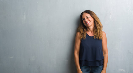 Middle age hispanic woman standing over grey grunge wall looking away to side with smile on face, natural expression. Laughing confident.