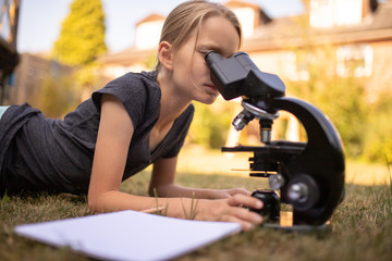 Ein 9 jähriges Mädchen liegt auf dem Rasen im Garten und schaut in das Okular eines Mikroskops. Vor ihr liegt ein Blatt Papier