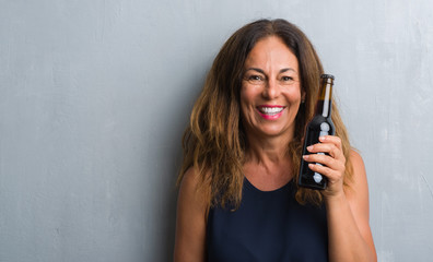 Middle age hispanic woman standing over grey grunge wall holding beer bottle with a happy face standing and smiling with a confident smile showing teeth