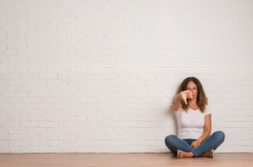 Middle age hispanic woman over white brick wall with angry face, negative sign showing dislike with thumbs down, rejection concept