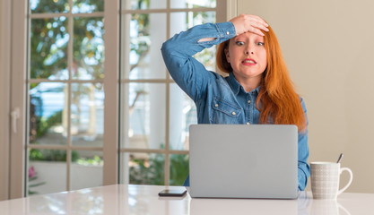 Redhead woman using computer laptop at home stressed with hand on head, shocked with shame and...