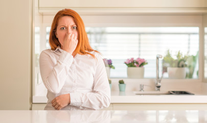Redhead woman at kitchen smelling something stinky and disgusting, intolerable smell, holding breath with fingers on nose. Bad smells concept. - obrazy, fototapety, plakaty