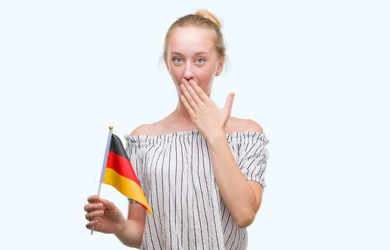 Blonde teenager woman holding flag of Germany cover mouth with hand shocked with shame for mistake, expression of fear, scared in silence, secret concept