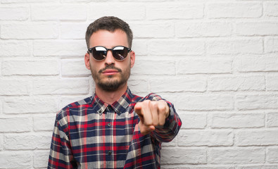 Young adult man wearing sunglasses standing over white brick wall pointing with finger to the...