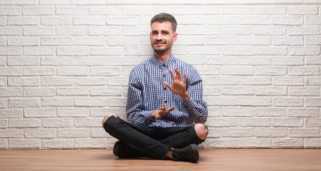 Young adult man sitting over white brick wall disgusted expression, displeased and fearful doing disgust face because aversion reaction. With hands raised. Annoying concept.