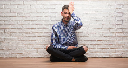 Young adult man sitting over white brick wall surprised with hand on head for mistake, remember...