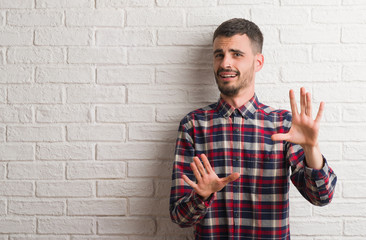 Young adult man standing over white brick wall afraid and terrified with fear expression stop...