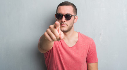 Young caucasian man over grey grunge wall wearing sunglasses pointing with finger to the camera and...
