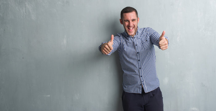 Young caucasian man over grey grunge wall approving doing positive gesture with hand, thumbs up smiling and happy for success. Looking at the camera, winner gesture.