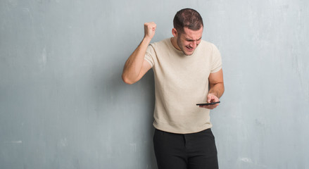 Young caucasian man over grey grunge wall texting a message using smartphone annoyed and frustrated shouting with anger, crazy and yelling with raised hand, anger concept