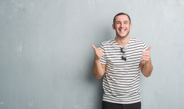 Young caucasian man over grey grunge wall success sign doing positive gesture with hand, thumbs up smiling and happy. Looking at the camera with cheerful expression, winner gesture.