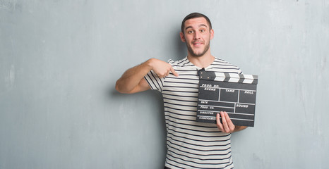 Young caucasian man over grey grunge wall holding movie clapboard with surprise face pointing...