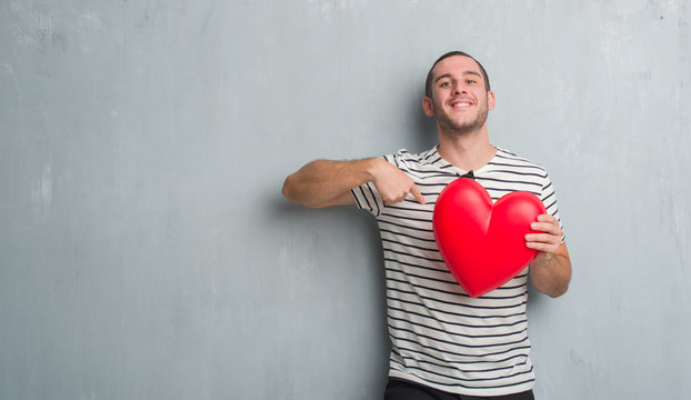 Young Caucasian Man Over Grey Grunge Wall Holding Red Heart With Surprise Face Pointing Finger To Himself