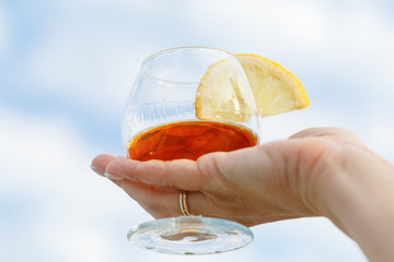 Goblet of cognac with a slice of lemon in a female hand against a blue sky background