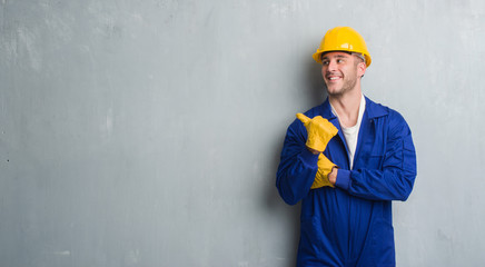 Young caucasian man over grey grunge wall wearing contractor uniform and safety helmet pointing and showing with thumb up to the side with happy face smiling - obrazy, fototapety, plakaty