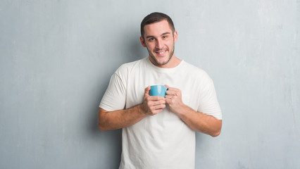Young caucasian man over grey grunge wall driking a cup of coffee with a happy face standing and...